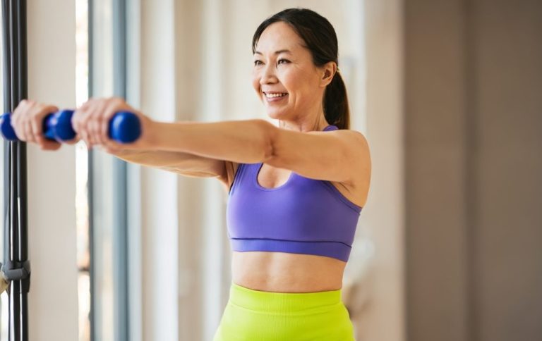 A woman smiles while exercising with blue dumbbells in a bright, spacious room.